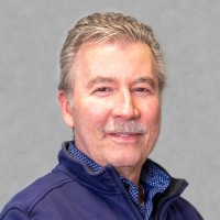 A middle-aged man with gray hair and a mustache, wearing a navy blue jacket over a blue collared shirt, poses in front of a plain light gray background, smiling softly at the camera.