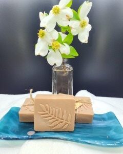 A 16 oz Apothecary Jar Candle in Lavender stands behind three beige soaps with leaf imprints, arranged on a blue ceramic tray over a white cloth.