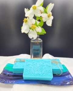 A 16 oz Apothecary Jar Candle in Lavender stands behind two ornate turquoise soaps on a blue and green glass dish with a white towel underneath, set against a dark background.