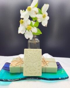A 16 oz Apothecary Jar Candle in Lavender sits behind intricately decorated white soap bars, all displayed on a turquoise and blue glass tray with a white cloth underneath.