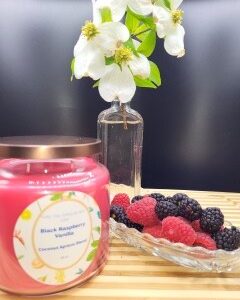 A lavender-scented candle in a 16 oz apothecary jar sits on a wooden surface beside a glass dish of fresh berries, with white flowers in a clear vase set against a dark background.