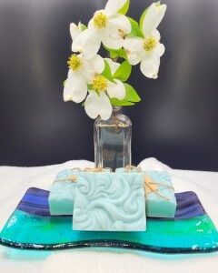 A 16 oz Apothecary Jar Candle in Lavender is displayed on a multicolored glass tray beside three light blue decorative soap bars, with white flowers in a glass vase, all set atop a white towel against a dark background.