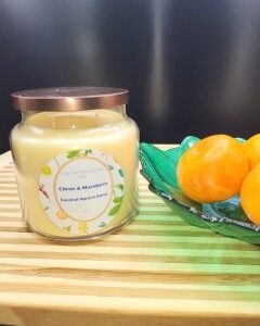 A 16 oz Apothecary Jar Candle labeled Lavender with a copper lid sits on a wooden striped surface next to a green dish of bright orange mandarins, set against a dark background.
