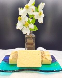 A small glass vase with white flowers is placed behind three beige soap bars on a blue and green glass dish, set against a dark background.