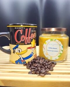 A Mickey Mouse-themed coffee mug, a 16 oz Apothecary Jar Candle - Lavender, and a small pile of coffee beans sit on a wooden surface with a dark background.