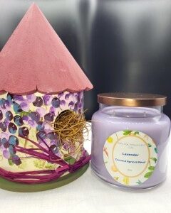 A colorful, hand-painted birdhouse with a pink roof sits next to a 16 oz Apothecary Jar Candle - Lavender in a glass jar with a golden lid and a floral label.