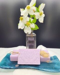 A 16 oz Apothecary Jar Candle - Lavender is displayed on a blue ceramic dish with a white cloth underneath, accompanied by a glass vase with white flowers behind it.