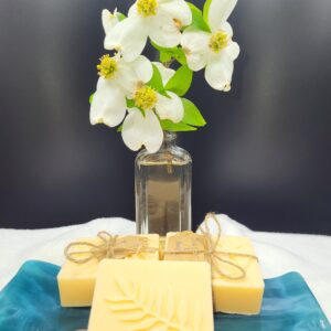 A 16 oz Apothecary Jar Candle in Lavender sits behind a blue ceramic tray with handmade yellow soap bars—one with a fern design, others tied with twine.