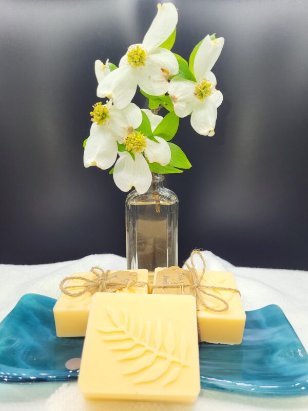 A 16 oz Apothecary Jar Candle in Lavender sits behind a blue ceramic tray with handmade yellow soap bars—one with a fern design, others tied with twine.