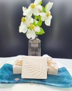 A 16 oz Apothecary Jar Candle in Lavender sits behind three soap bars, one with a fern leaf design, arranged on a blue ceramic dish. The dark background creates a clean, minimalistic setting.