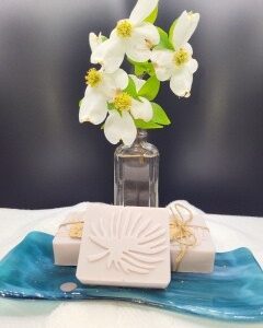 A 16 oz Apothecary Jar Candle in Lavender sits behind two wrapped soaps and a leaf-design soap, all arranged on a blue ceramic dish atop a white surface.