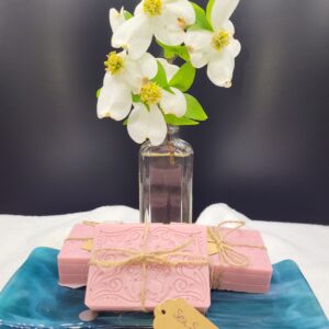 A 16 oz Apothecary Jar Candle in Lavender stands behind three pink ornate soaps tied with twine, resting on a blue dish. A tag labeled Sea Salt & Orchid lies in front of the display.
