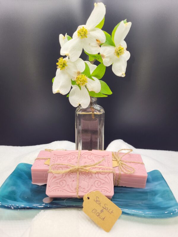 A 16 oz Apothecary Jar Candle in Lavender stands behind three pink ornate soaps tied with twine, resting on a blue dish. A tag labeled Sea Salt & Orchid lies in front of the display.