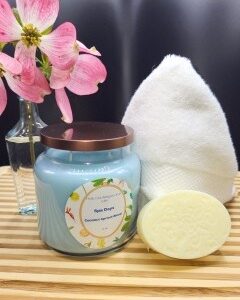 A 16 oz Apothecary Jar Candle - Lavender, a white folded towel, an oval bar of soap, and a vase with pink flowers are arranged on a wooden surface against a dark background.