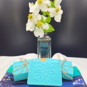 A 16 oz Apothecary Jar Candle - Lavender stands behind a blue ceramic tray holding three decorative blue soaps tied with twine, each featuring an ornate, embossed floral pattern.