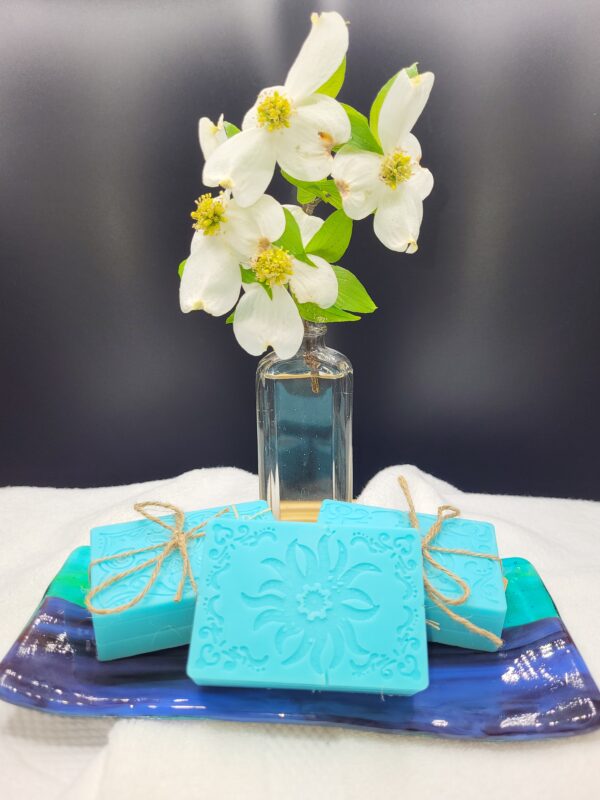 A 16 oz Apothecary Jar Candle - Lavender stands behind a blue ceramic tray holding three decorative blue soaps tied with twine, each featuring an ornate, embossed floral pattern.