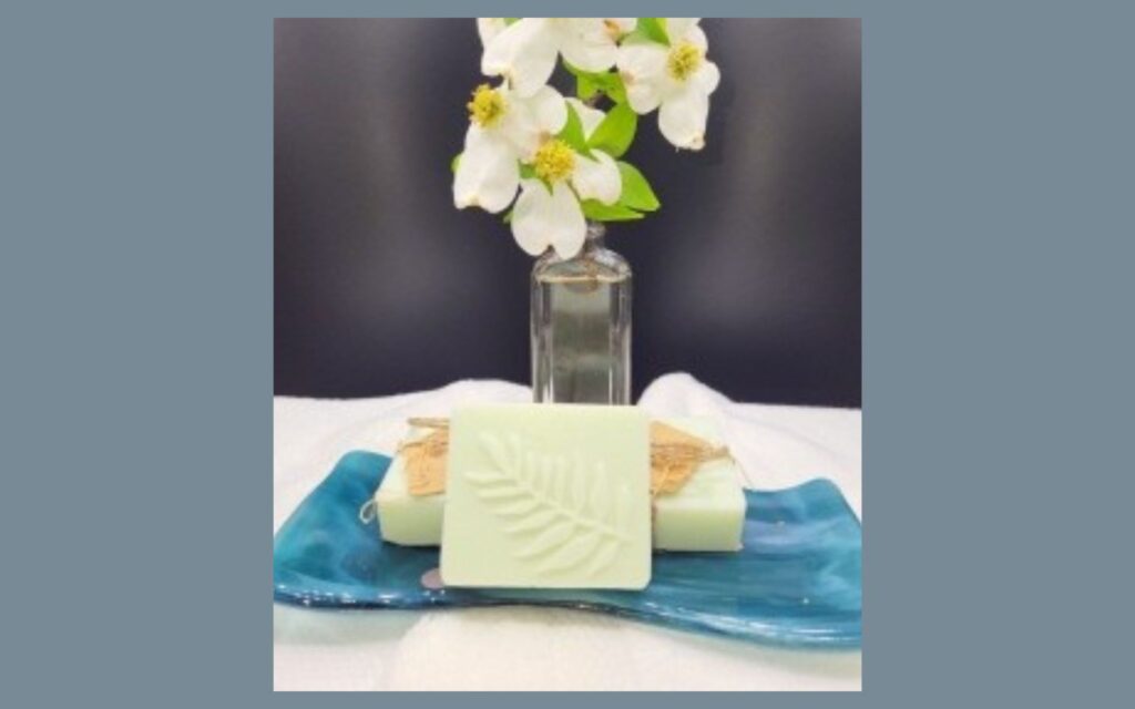 A bar of light green soap with a leaf imprint sits on a blue ceramic dish, with a glass vase of white flowers and green leaves in the background.