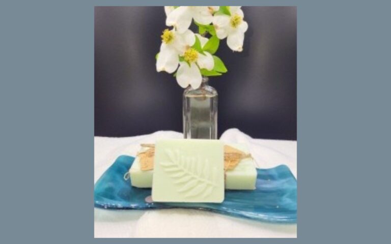 A bar of light green soap with a leaf imprint sits on a blue ceramic dish, with a glass vase of white flowers and green leaves in the background.