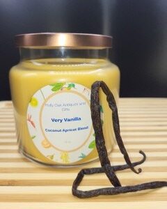 A 16 oz Apothecary Jar Candle - Lavender sits on a wooden surface with two dried vanilla beans in front. The candle is a soft lavender color and features a copper-colored lid.