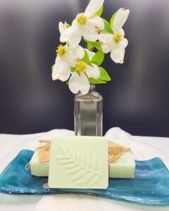 A 16 oz Apothecary Jar Candle in Lavender sits behind a blue dish with two pale green soap bars, one adorned with a fern leaf design. The dark background contrasts with the light-colored items.