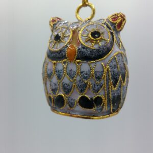 A decorative owl ornament with gold detailing hangs from a gold string against a plain light background. The owl features intricate patterns and has black, gray, and gold coloring.