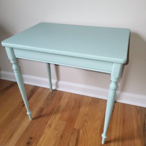 A small, light blue wooden table with four slender, turned legs stands on a hardwood floor against a light-colored wall.
