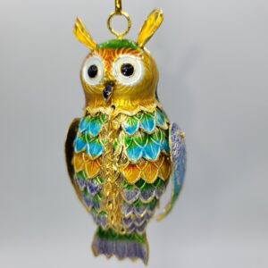 A colorful owl ornament with gold, green, blue, and purple details hangs from a thin gold string against a plain light background.
