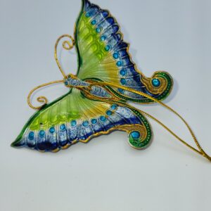 A decorative butterfly ornament with vibrant green, blue, and yellow wings, detailed with gold trim and blue dots, set against a plain white background. Thin gold wires extend from its body, mimicking antennae and tail.