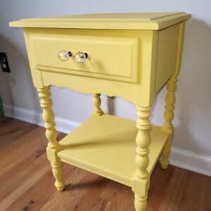 A yellow wooden nightstand with ornate legs, a single drawer featuring two glass knobs, and a lower shelf, placed on a hardwood floor against a light-colored wall.