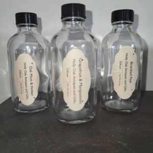 Three clear glass bottles with black caps, each labeled with different scents: Oak Moss & Amber, Grapefruit & Mangosteen, and Bourbonized Pear, on a dark surface against a light wall.