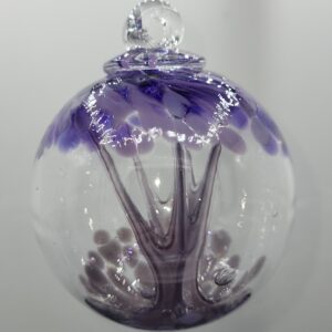 A clear glass ornament with a tree design inside, featuring a purple trunk and branches with lavender and white leaf-like details, hanging against a plain, light background.