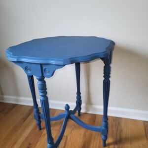 A small, ornate side table painted matte blue stands on a hardwood floor against a plain white wall. The table has decorative legs and a scalloped edge on its top.