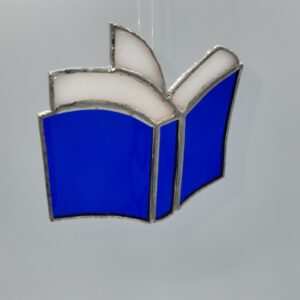 A stained glass ornament shaped like an open book, with blue pages and white edges outlined in silver, hanging against a gray background.