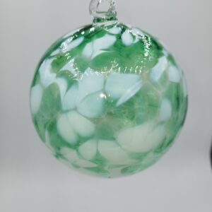 A round glass ornament with a loop on top, featuring a swirled pattern of green and white patches on a translucent background, hanging against a plain light gray backdrop.