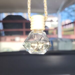 A close-up of a diamond-shaped glass car air freshener with a wooden cap, hanging by a string inside a car. The background is blurred, showing part of the dashboard, a grill, and outdoor scenery.