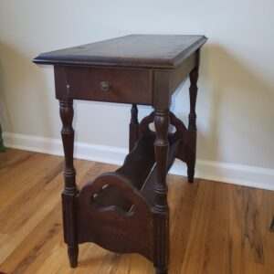 A vintage wooden side table with a single drawer and an open magazine rack underneath, standing on hardwood flooring near a light-colored wall and a patterned rug.
