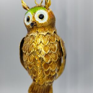 A close-up of a shiny, gold-colored owl ornament with green and white around its eyes, hanging by a gold string against a blurred background.