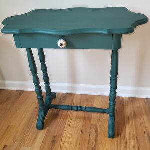 A small, vintage-style teal accent table with a scalloped edge top, a single drawer featuring a decorative white knob, and turned legs, standing on a hardwood floor against a light-colored wall.