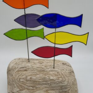 A wooden base holds six colorful stained glass fish—orange, purple, blue, green, yellow, and red—mounted on thin metal rods, arranged in two rows against a plain white background.