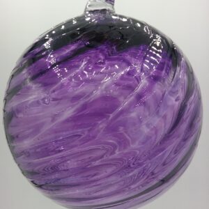 A round, hand-blown glass ornament with swirling patterns of purple, black, and clear glass, hanging against a plain white background.