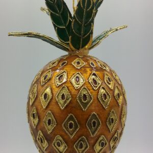 A decorative ornament shaped like a pineapple, adorned with gold and brown embroidery and green, gold-trimmed leaves at the top, hangs from a gold string against a plain background.
