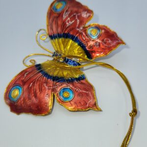 A decorative butterfly ornament with red, yellow, and blue patterns on its wings, outlined in gold, and a gold string attached, displayed on a white surface.