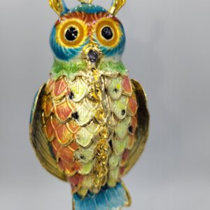 A close-up of a colorful, ornate owl ornament with gold accents, textured feathers in shades of green, red, blue, and yellow, and large, round eyes, hanging against a plain light background.