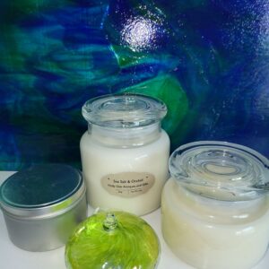 Three candles in glass jars and a silver tin are displayed in front of an abstract blue and green background. A small, round, green glass ornament sits in front of the candles.