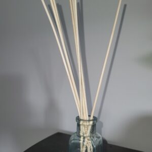 A clear glass bottle holds several long, thin wooden sticks, arranged in a fan shape. The bottle sits on a dark surface against a plain, light gray background.