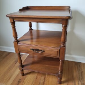 A wooden side table with turned legs, a single drawer with a brass handle in the center, an open middle shelf, and a lower shelf, set on a hardwood floor against a white wall.