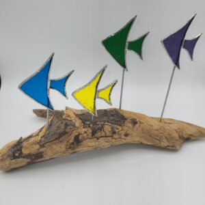 Four stained glass fish in blue, yellow, green, and purple are mounted on metal rods attached to a piece of driftwood, set against a plain white background.