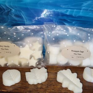 Two bags of white wax melts labeled Very Vanilla and Pineapple Sage sit on a wooden surface. Three leaf-shaped wax melts are displayed in front. A blue, swirling background is behind the bags.
